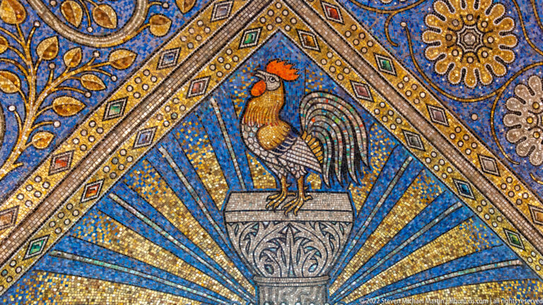 Colorful rooster standing on a pedestal within a triangular blue-and-gold radiating mosaic panel.