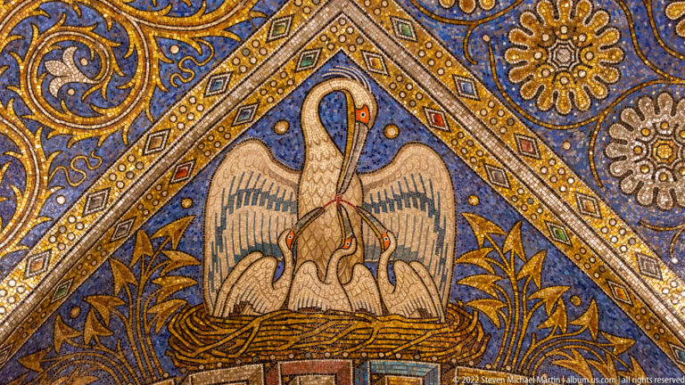 Pelican bending over chicks in a nest within a blue-and-gold triangular mosaic panel.