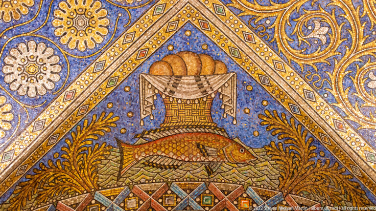 Covered vessel or basket with rounded loaves above a large fish in a blue-and-gold mosaic panel.