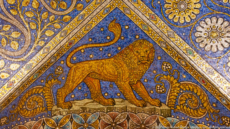 Golden lion in profile with raised curling tail on a blue mosaic ground framed by rosettes and gold ornament.
