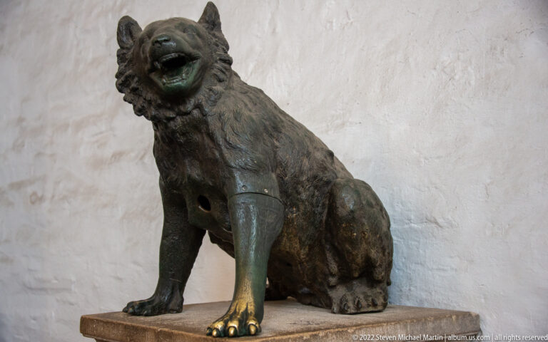 Seated dark bronze animal with raised head, open mouth, and green patina on its legs against a pale wall.