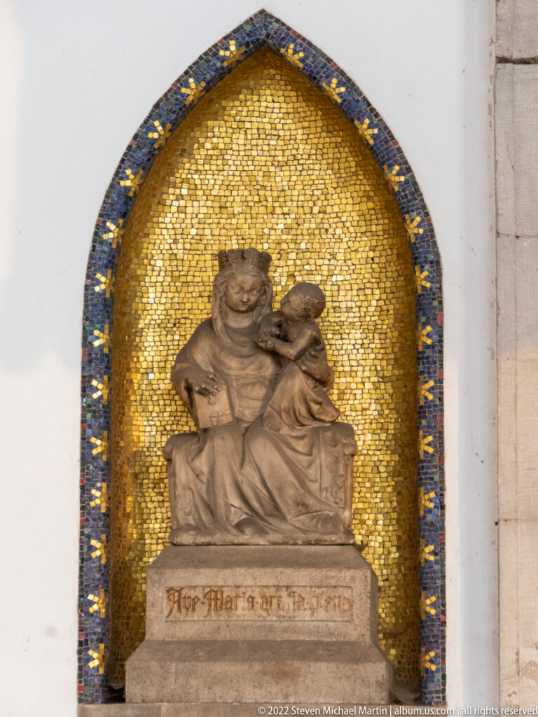 Brown figural sculpture of an adult holding a child set in a pointed niche lined with gold mosaic.
