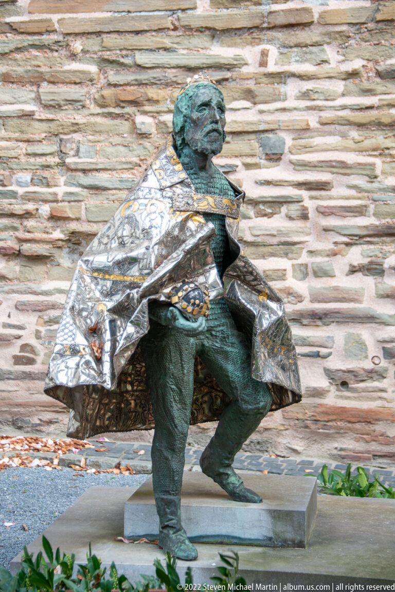 Seated bronze male figure with dark patina and a bright faceted cloak against a rough stone wall.