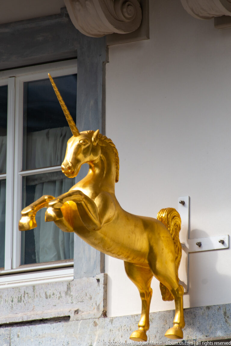 Gilded unicorn rearing beneath a covered façade opening, with one foreleg raised and horn angled upward.