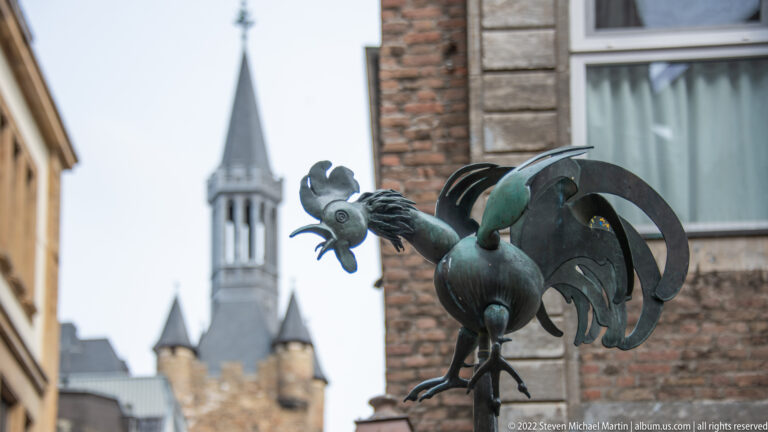 Metal rooster mounted on an ornamental bracket, with a church or civic tower blurred in the background.