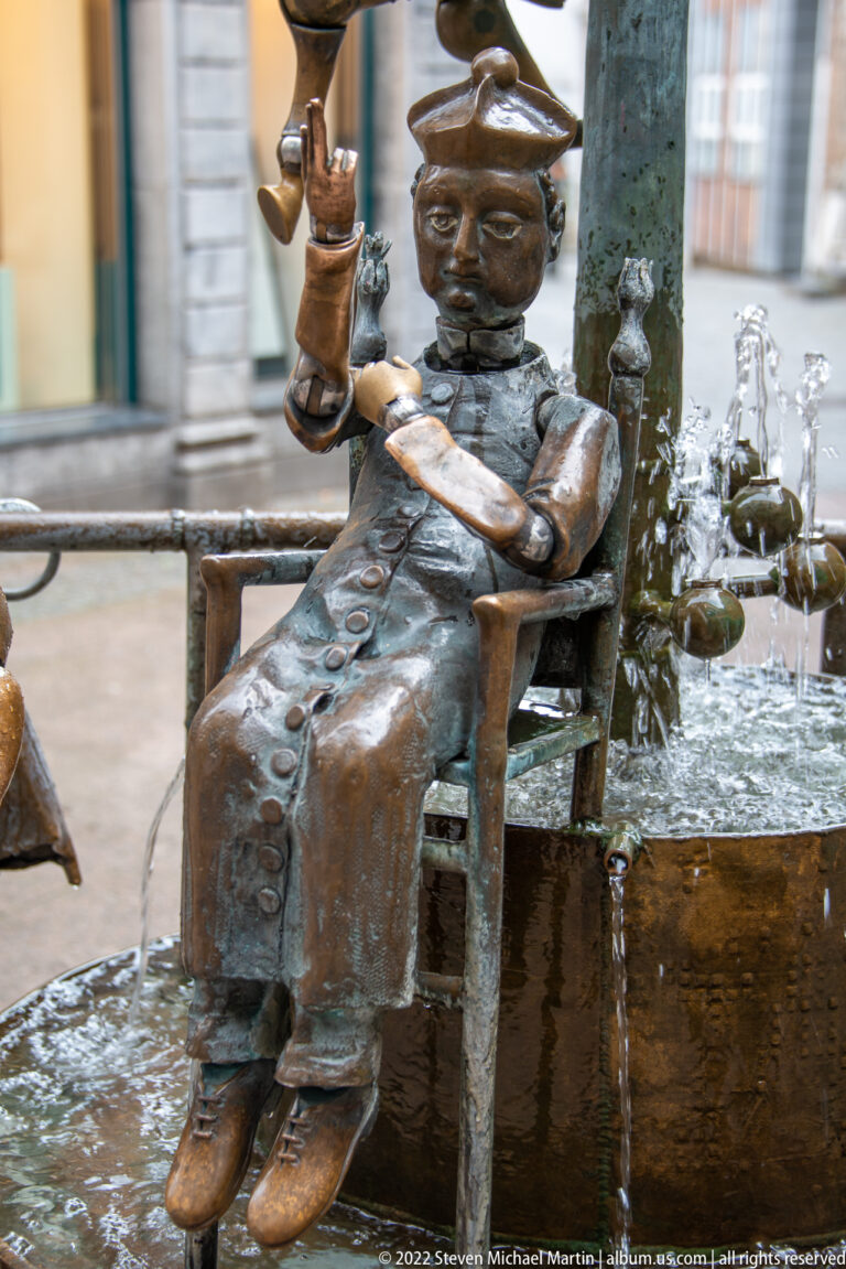 Bronze seated woman in a hat beside a fountain, with water jets and small sculptural figures behind her.