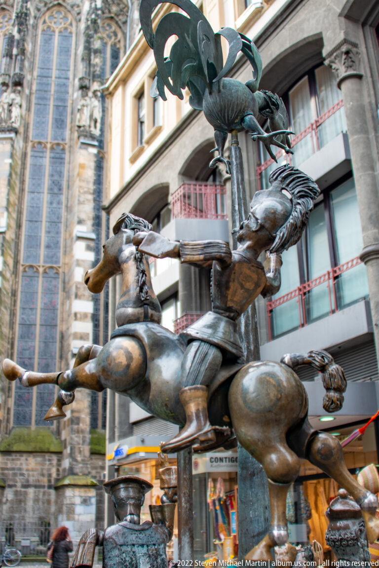 Clustered bronze fountain figures with people and rearing animal forms, surrounded by historic buildings.