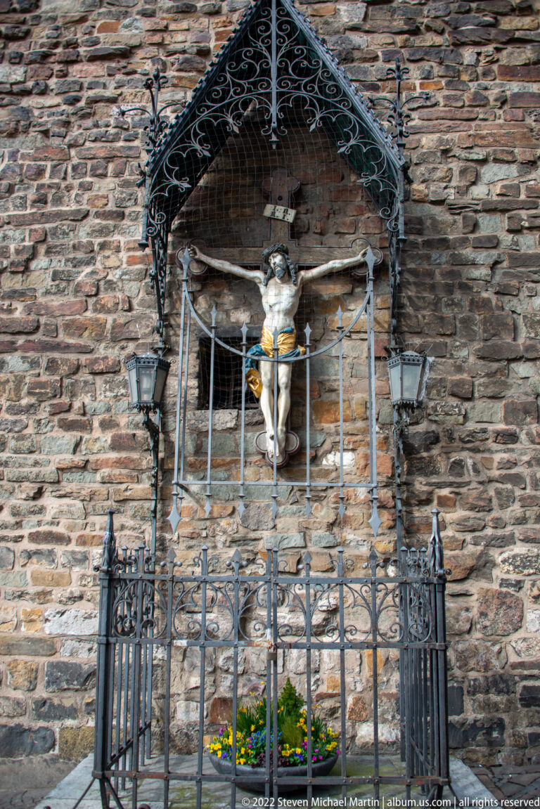 Crucifix shrine beneath an ornate metal canopy, enclosed by iron railings and flanked by wall lamps.