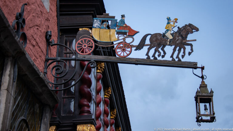 Painted hanging sign of a horse-drawn coach on a wrought bracket, with a small lantern suspended below.