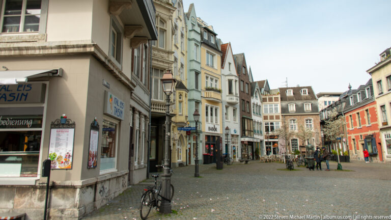 Pedestrian street lined with narrow townhouses, shopfronts, bicycles, and a small square in central Aachen.
