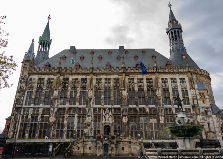 Broad façade of Aachen Town Hall with tall windows, statues, steep roof, and towers under a cloudy sky.