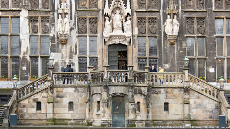 Stone staircase and carved entrance of Aachen Town Hall, framed by tall windows, tracery, and statues.