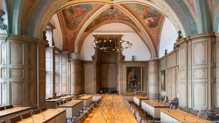 Long paneled hall with benches, ribbed vaults, painted ceiling scenes, and a large circular chandelier.