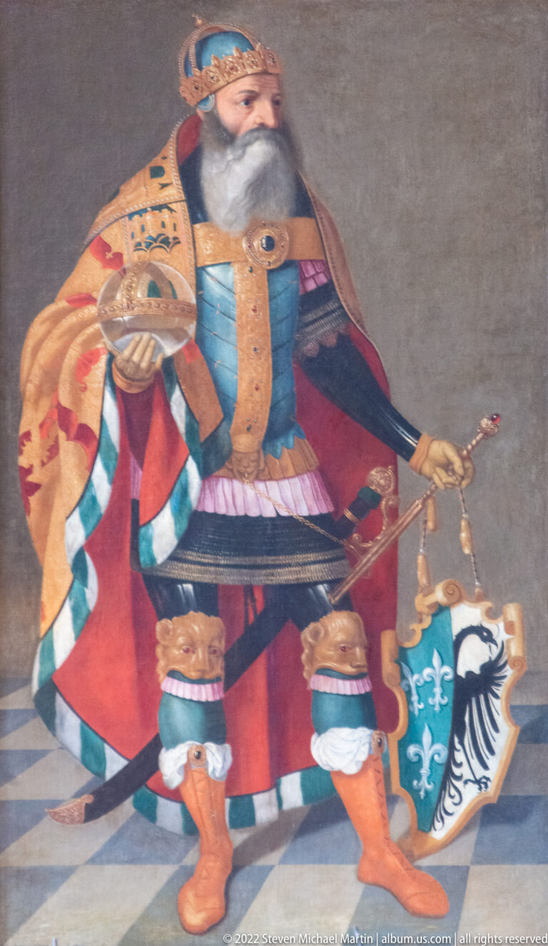 Painted bearded king wearing a crown and ornate robes, holding a scepter beside a heraldic shield.