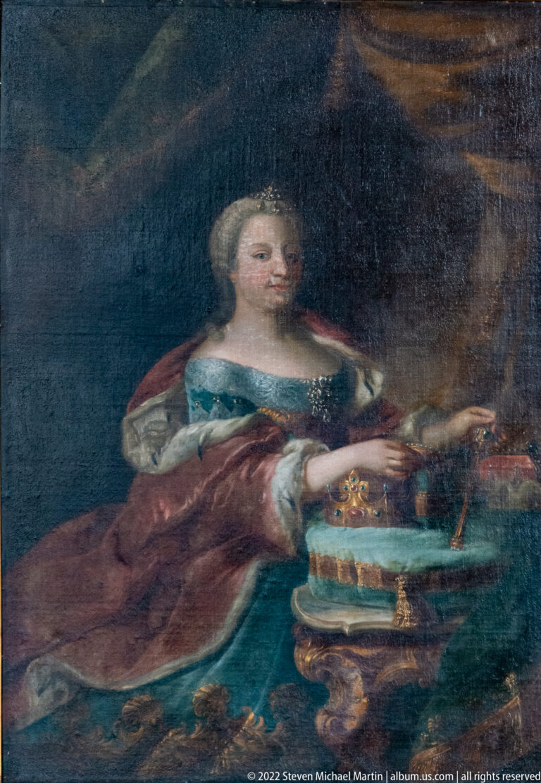 Formal painted portrait of an aristocratic woman in a rich gown and mantle seated beside a table.