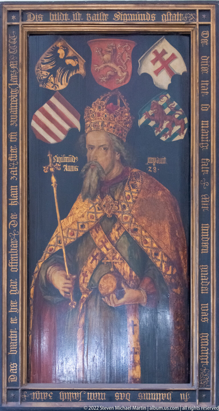 Painted crowned ruler in ornate robes surrounded by heraldic shields and inscriptions.