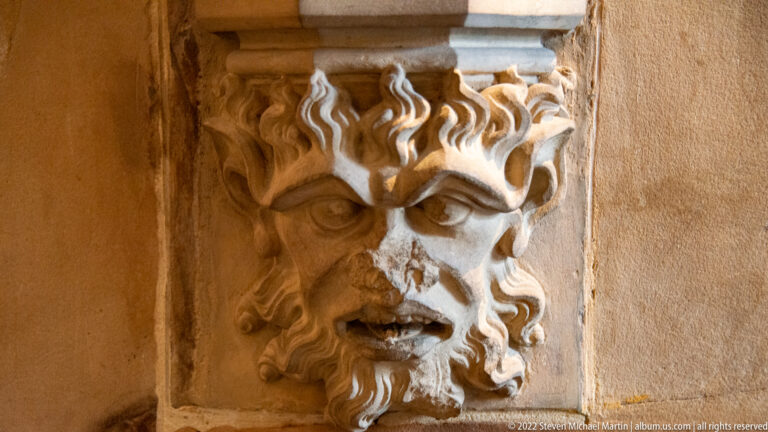 Close view of a carved stone grotesque head with deep brows, curling hair, and open mouth on an architectural corner.