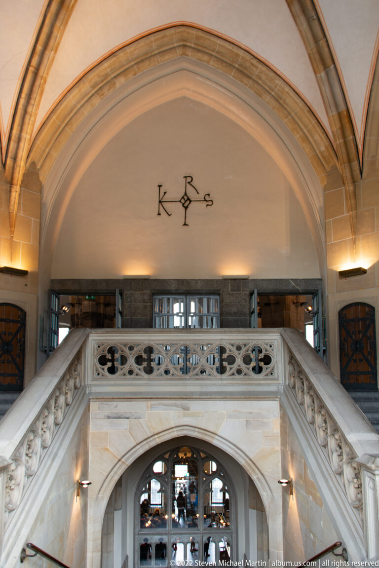 Broad stone staircase with carved balustrade beneath pointed vaulting and a monogram in Aachen Town Hall.