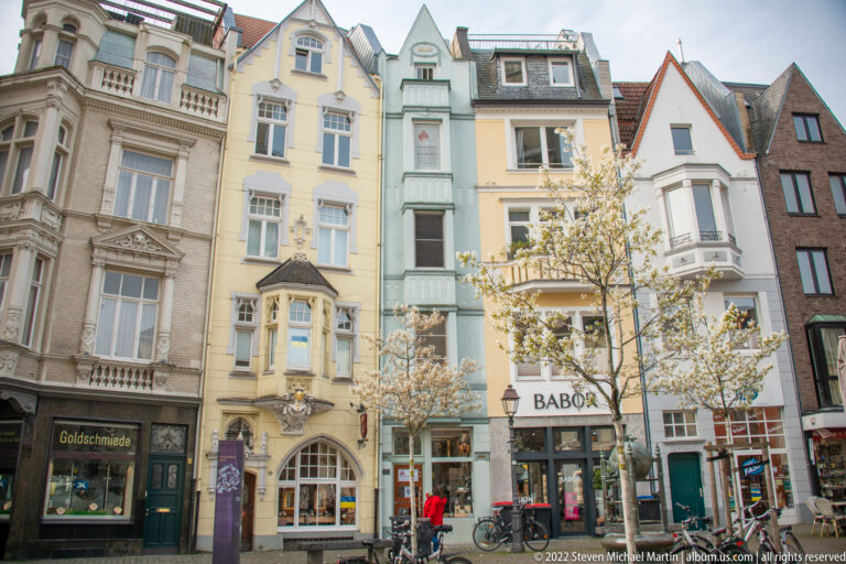 Row of narrow historic townhouses with ornate windows, bay projections, shops, bicycles, and small street trees.
