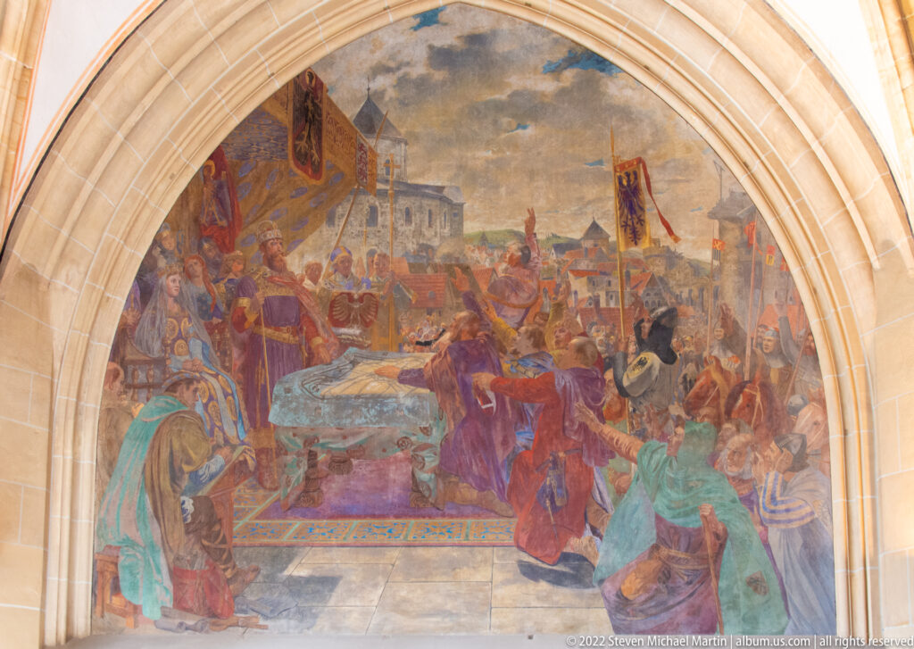 Large arched mural of a crowded royal ceremony with banners, clerics, soldiers, and richly dressed figures.