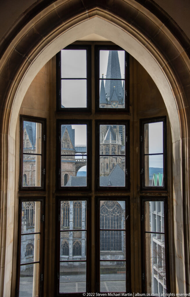 Large arched window with dark mullions framing Aachen Cathedral and neighboring historic roofs.