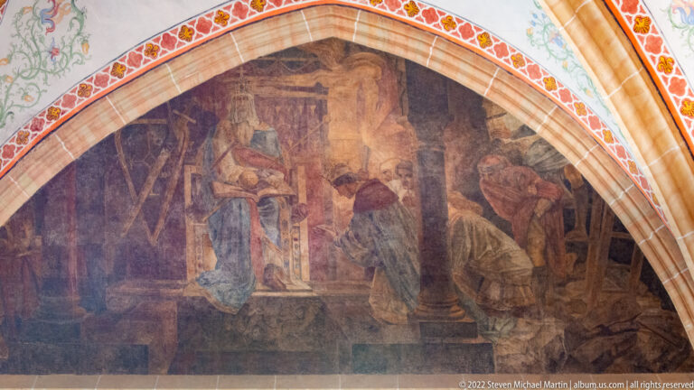Arched mural with robed men gathered around a seated figure in an architectural interior.