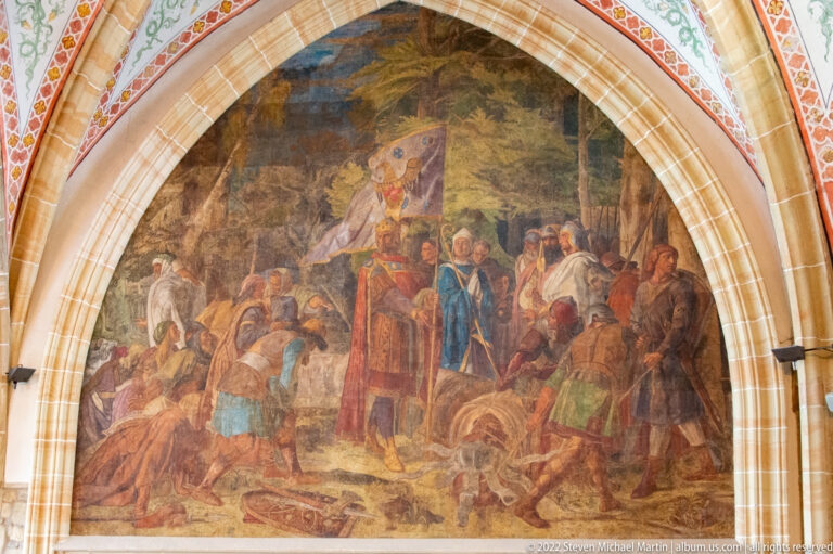 Arched mural showing a red-robed central figure addressing a crowd of soldiers and attendants.