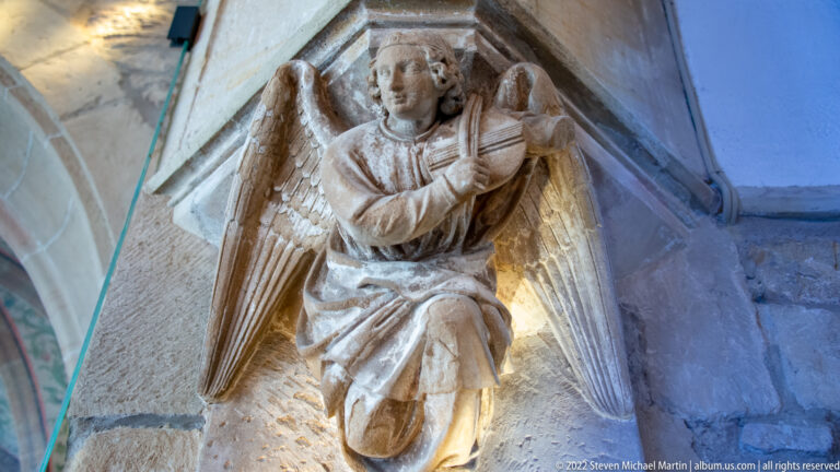 Carved stone angel with spread wings kneeling beneath a vault or architectural support.