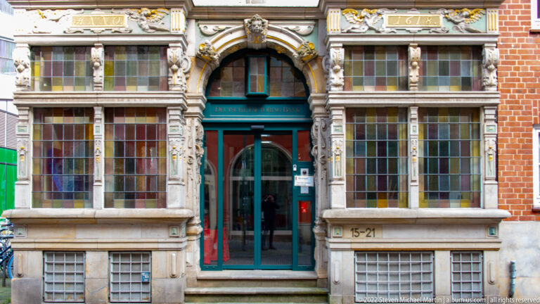 Ornate brick-and-stone façade with leaded-glass windows, sculpted figures, gilded details, and a central arched doorway.