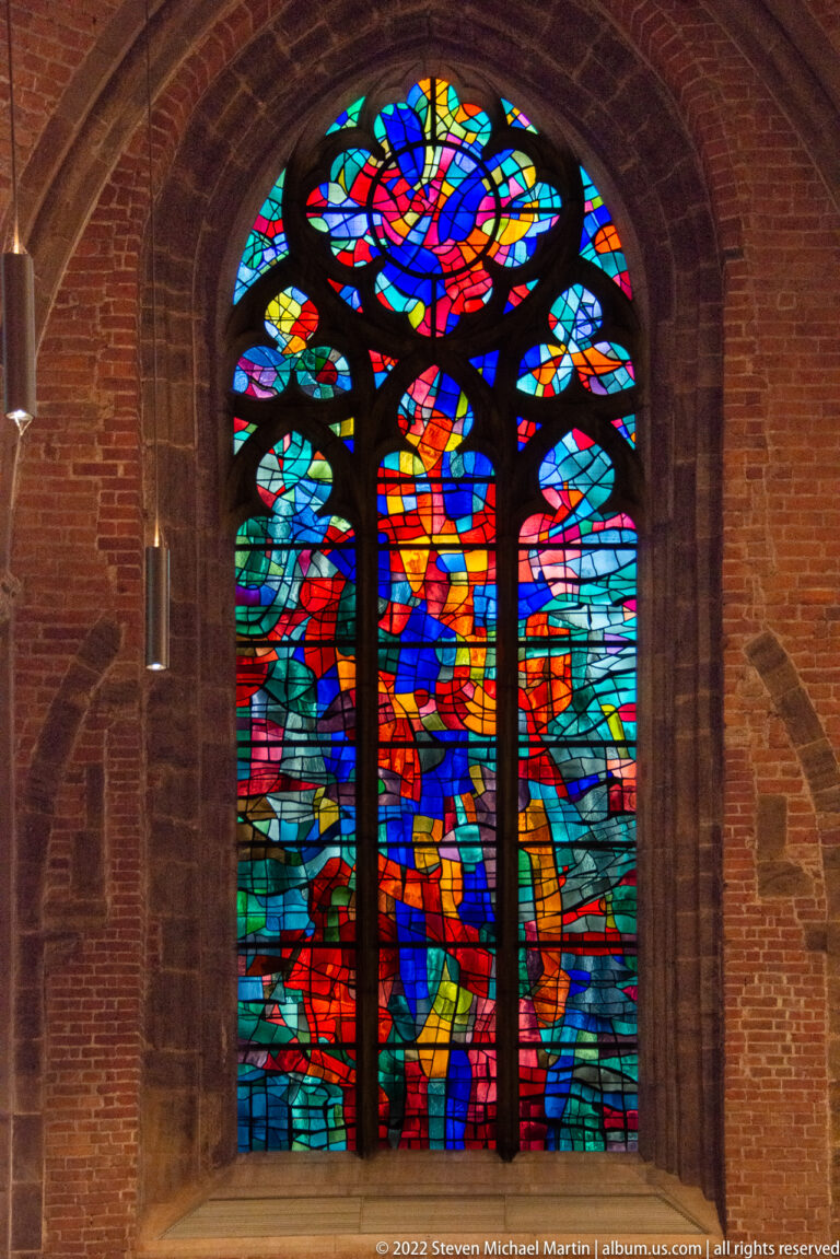 Abstract multicolored stained-glass window set in a tall pointed opening within a red-brick church wall.