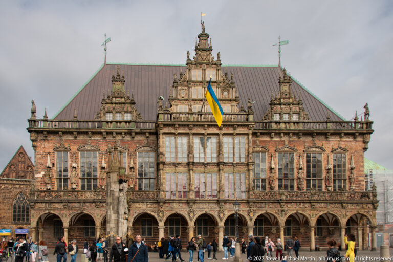 Full frontal view of Bremen Town Hall with arcades, ornate gables, steep roof, pedestrians, and a Ukrainian flag.
