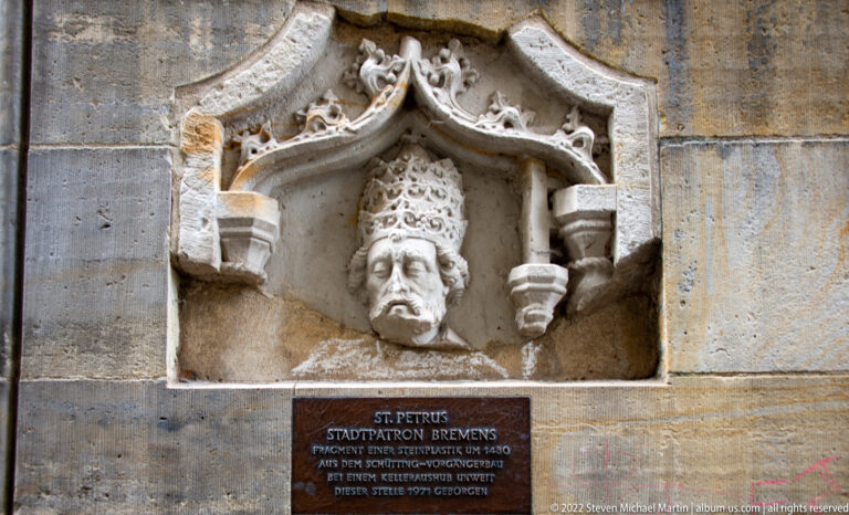 Stone head of Saint Peter wearing a papal tiara in a small Gothic niche above an identifying plaque.