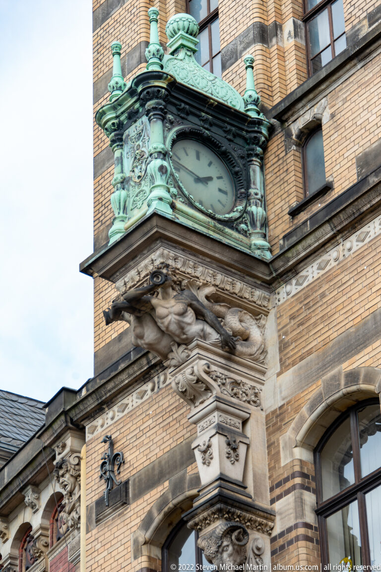Large green-patinated façade clock surrounded by carved stone ornament and patterned brickwork.