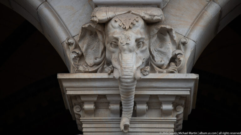 Stone elephant head with long trunk carved into the capital beneath an architectural arch.
