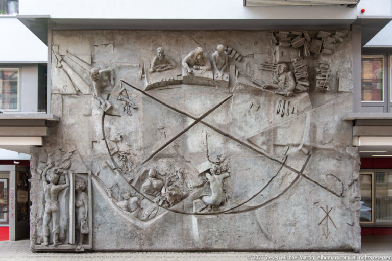 Large modern stone wall relief with multiple human figures, objects, and sweeping incised diagonal lines.