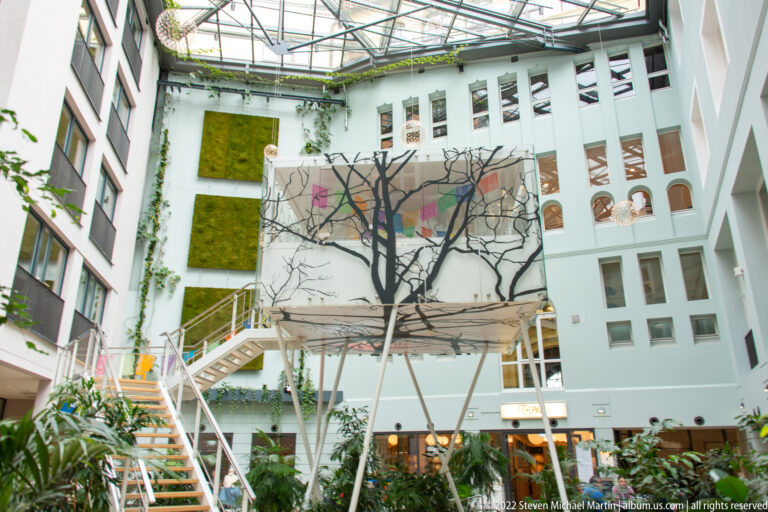 Glass-roofed interior atrium with plants, staircases, green wall panels, and a suspended black-tree artwork.