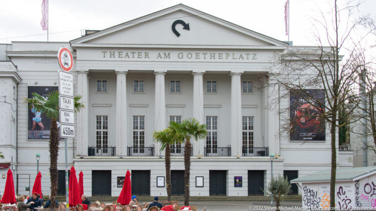 White neoclassical Theater am Goetheplatz with tall columns, triangular pediment, palm trees, and performance posters.