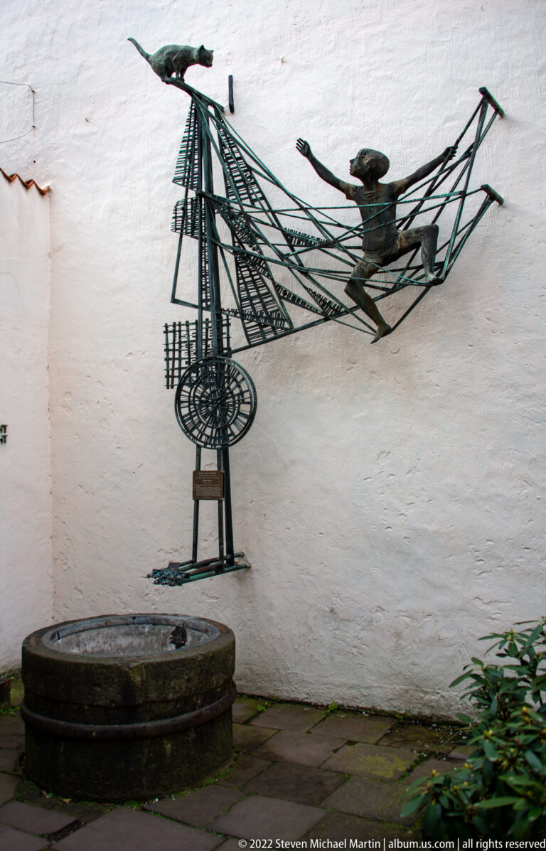 Dark metal wall sculpture of a human figure within an elongated framework above a low circular stone well.
