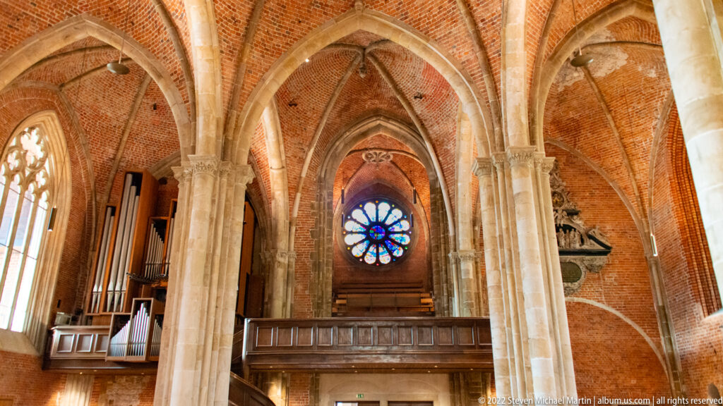 Pipe organ on a wooden gallery beneath brick vaults, with a circular stained-glass window behind it.