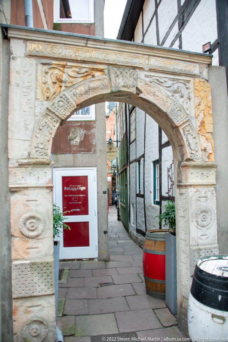 Decorative pale-stone arch opening onto a narrow historic lane lined with half-timbered and plastered buildings.
