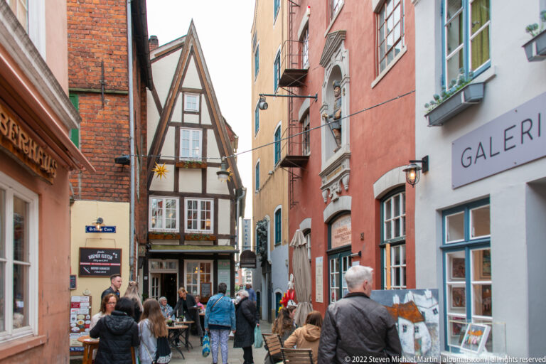 Narrow historic pedestrian street with colorful houses, half-timbering, café seating, gallery windows, and visitors.