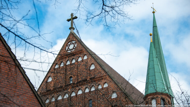 Brick church gable with clock, decorative masonry, cross, and tall green copper spire against a blue sky.