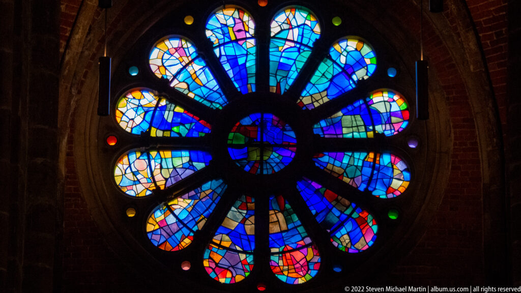 Round abstract stained-glass rose window with radiating blue, turquoise, yellow, red, and violet segments.