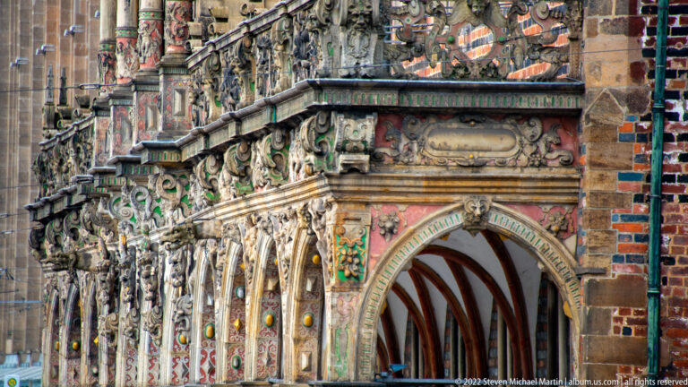Close oblique view of Bremen Town Hall’s ornate arcades with carved figures, heraldry, gilding, and painted decoration.