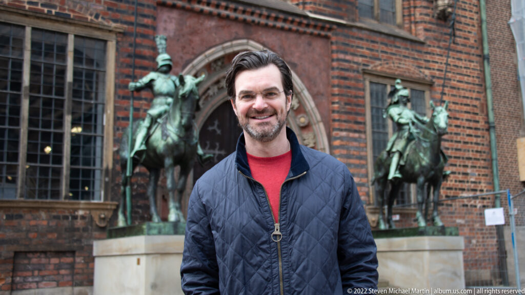 Smiling dark-haired man in a navy jacket standing before equestrian sculptures and a brick Gothic doorway in Bremen.