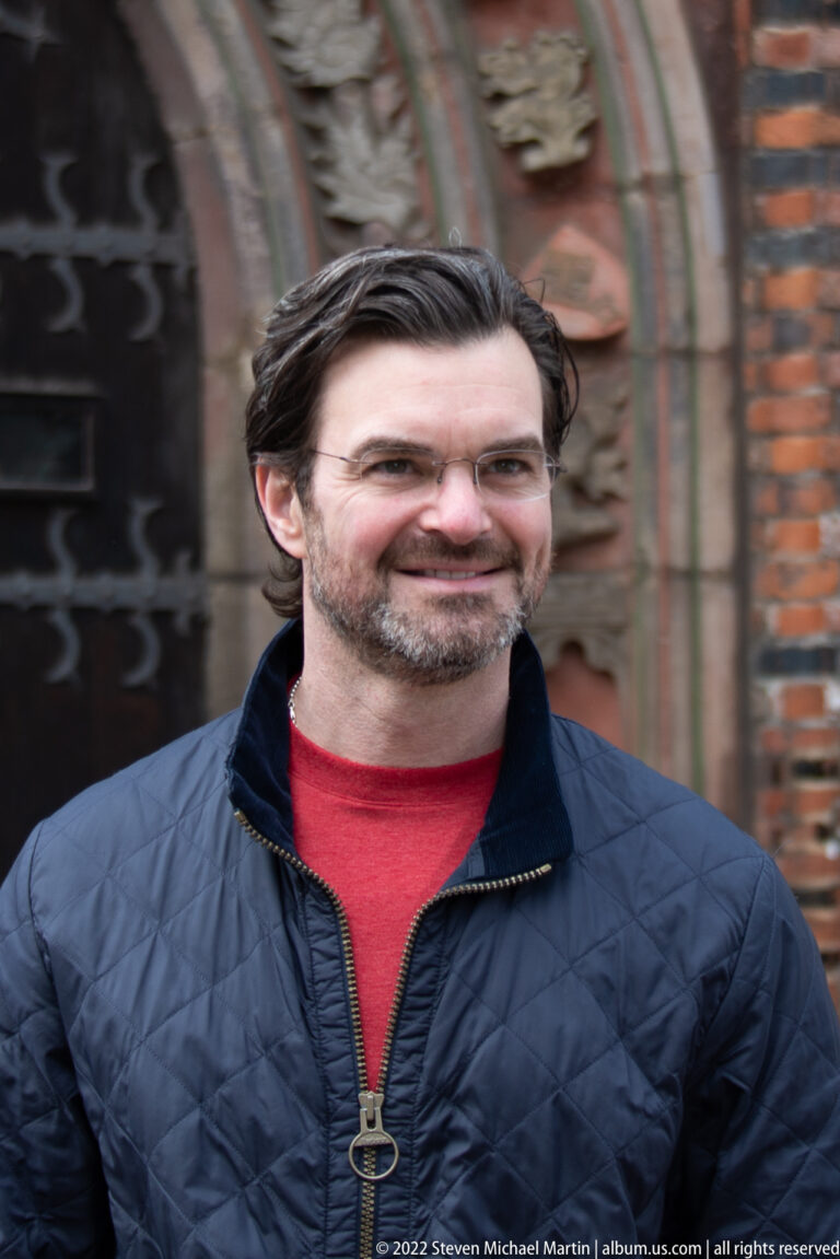 Close portrait of a smiling dark-haired man in a navy jacket and red shirt against blurred historic architecture.