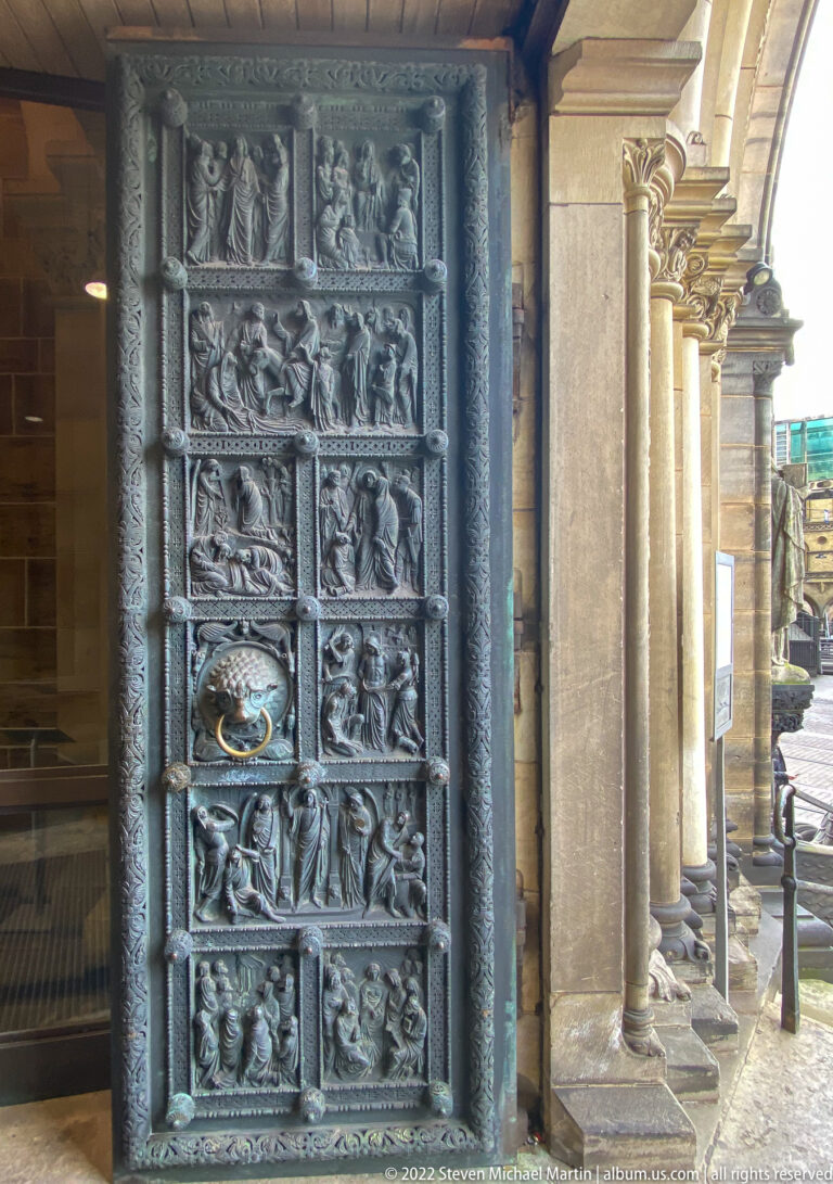 Dark bronze cathedral door with stacked biblical relief panels, ornate borders, and a projecting lion-head ring handle.