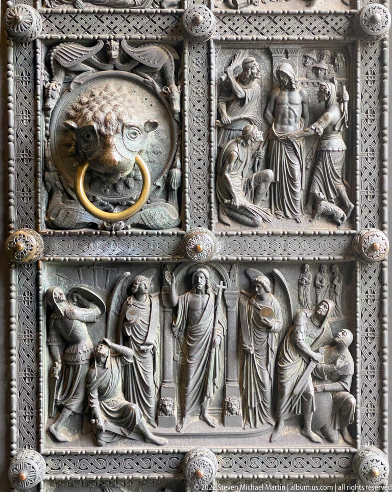Close view of bronze biblical relief panels surrounding a lion-head ring handle on a cathedral door.