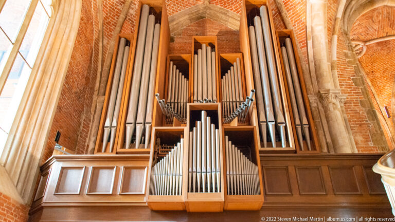 Large pipe organ with symmetrical metal pipes and wooden case inside a brick-and-stone Gothic church.
