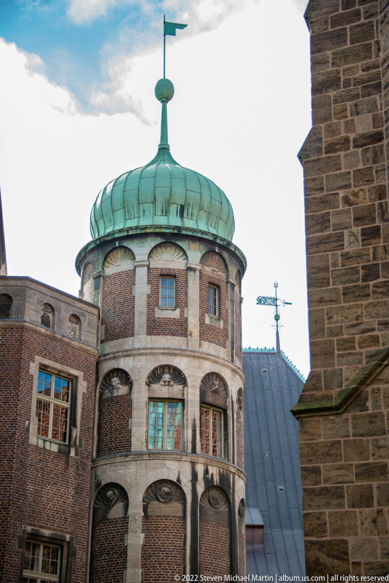 Round brick tower with green copper roof and weather vane, partly framed by neighboring historic buildings.